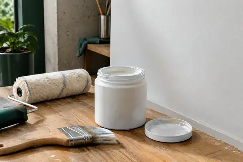Pot ouvert de peinture blanche satinée près d’un rouleau et d’un pinceau dans un atelier soigné.